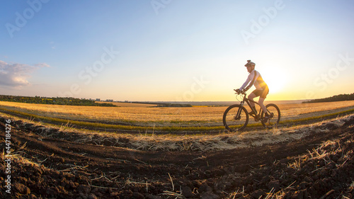 Fototapeta Naklejka Na Ścianę i Meble -  The cyclist rides a bike on the road near the field against the backdrop of the setting sun. Outdoor sports. Healthy lifestyle.