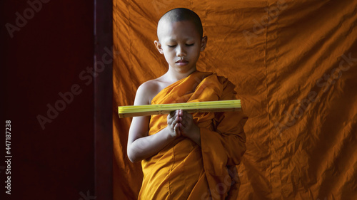 Fotografi portrait of Thai buddhist novice in orange robe standing and reading buddhism lessen scripture by window in temple
