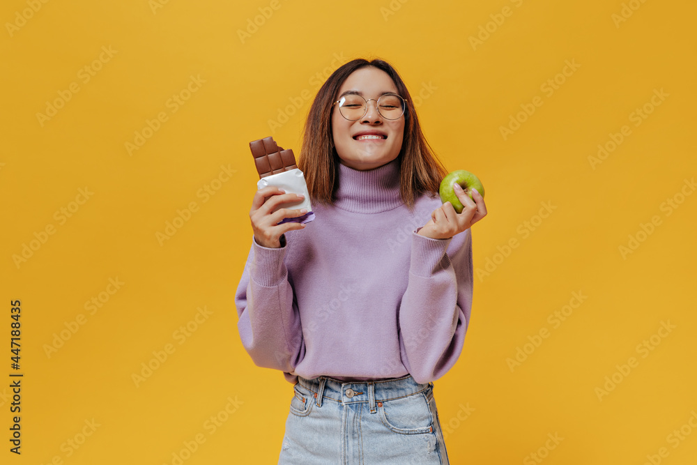 Happy young girl in eyeglasses smiles with closed eyes. Cheerful woman in purple sweater holds fresh green apple and tasty sweet milk chocolate bar on orange background. Happy young girl in eyeglasses smiles with closed eyes. Cheerful woman in purple sweater holds fresh green apple and tasty sweet milk chocolate bar on orange background.