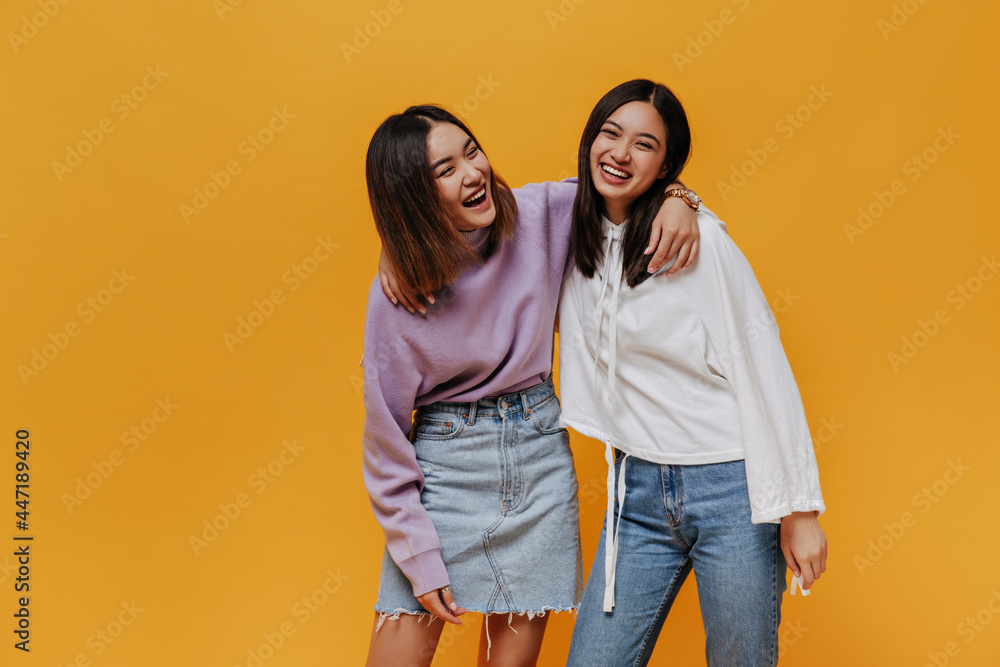 Joyful happy emotional girls laugh on orange background. Pretty Asian woman in denim skirt and purple sweater hugs friends on isolated.