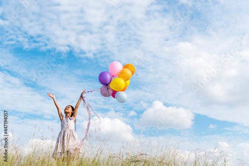 Fotografia Cheerful cute girl holding balloons running on green meadow white cloud and blue sky with happiness
