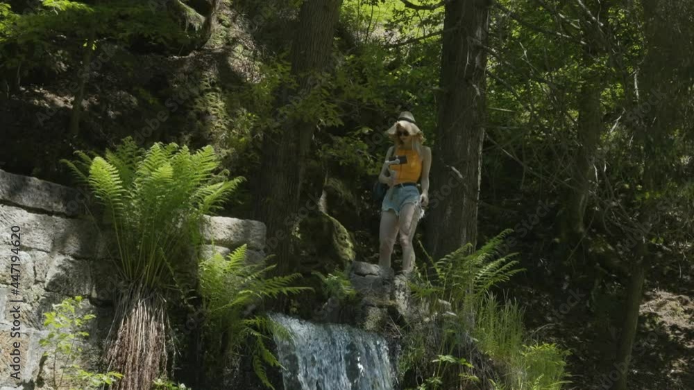 Young Blonde Girl With Floppy Hat Standing on Top of Waterfall and Recording Video With Smartphone and Gimbal, Slow Motion Young Blonde Girl With Floppy Hat Standing on Top of Waterfall and Recording Video With Smartphone and Gimbal, Slow Motion