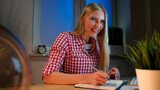 Lively young blond woman in casual checkered shirt, sitting at wooden desk with notebook holding metal pen and looking at camera smiling at night in dark room.