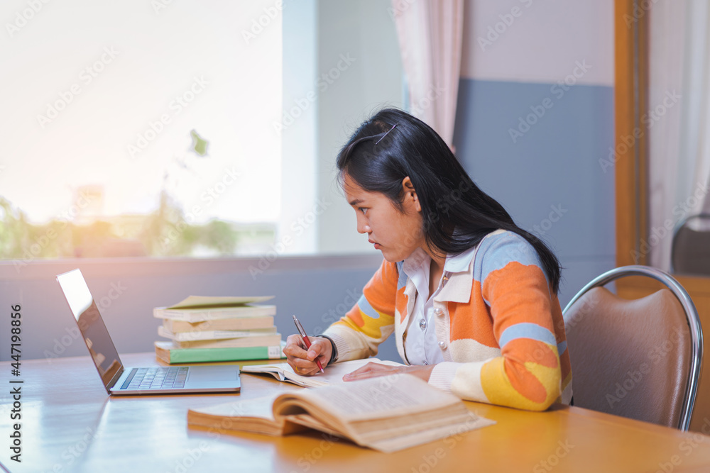 A stressful Asian college woman student in student uniform studying online, reading a book, using digital tablet or laptop in university library in free time by oneself A stressful Asian college woman student in student uniform studying online, reading a book, using digital tablet or laptop in university library in free time by oneself