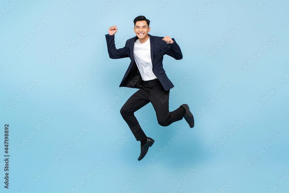 Full lenght portrait of ecstatic handsome Asian man jumping and raising his fists on isolated light blue studio background Full lenght portrait of ecstatic handsome Asian man jumping and raising his fists on isolated light blue studio background