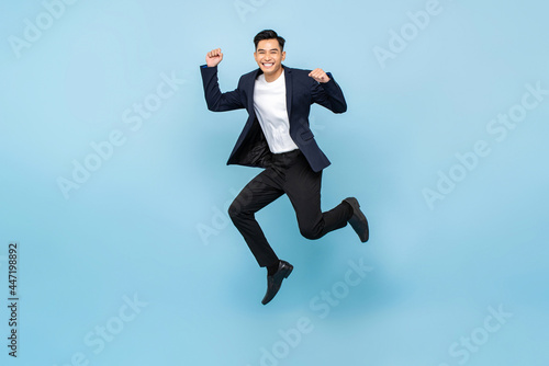 Full lenght portrait of ecstatic handsome Asian man jumping and raising his fists on isolated light blue studio background