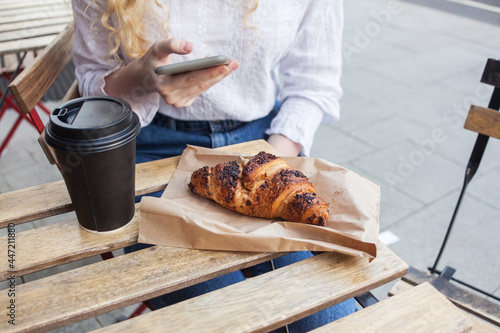 millennial hipster white girl texting on smart phone in a restaurant cafe terrace. cup of coffee to go and french croissant, quick snack, city street breakfast or brunch. unfocused background