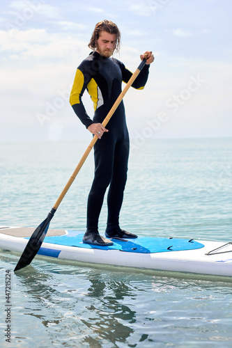 Bearded man with long hair in black wetsuit working out with stand up paddle board on water or in open sea,athelte caucasian guy is engaged in water sport, extreme sport. subsurfing concept