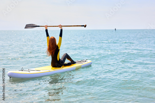 Rear view on redhead female in wetsuit raising paddle while surfing, enjoy active beach lifestyle, fit sporty caucasian lady engaged in standup paddleboarding