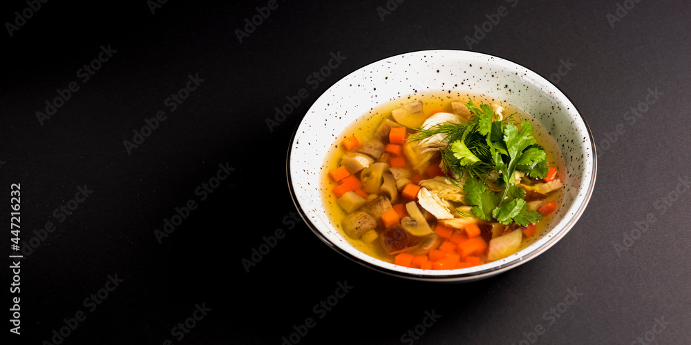 Chicken soup with vegetables amd mushrooms, fresh parsley and dill on top. Served in a white bowl over black background