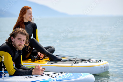 Sportive caucasian couple, young man and woman in wetsuit take a break after standup paddleboarding together, have rest at sea. Summer, vacation, sea, healthy lifestyle concept. focus on bearded man © Roman