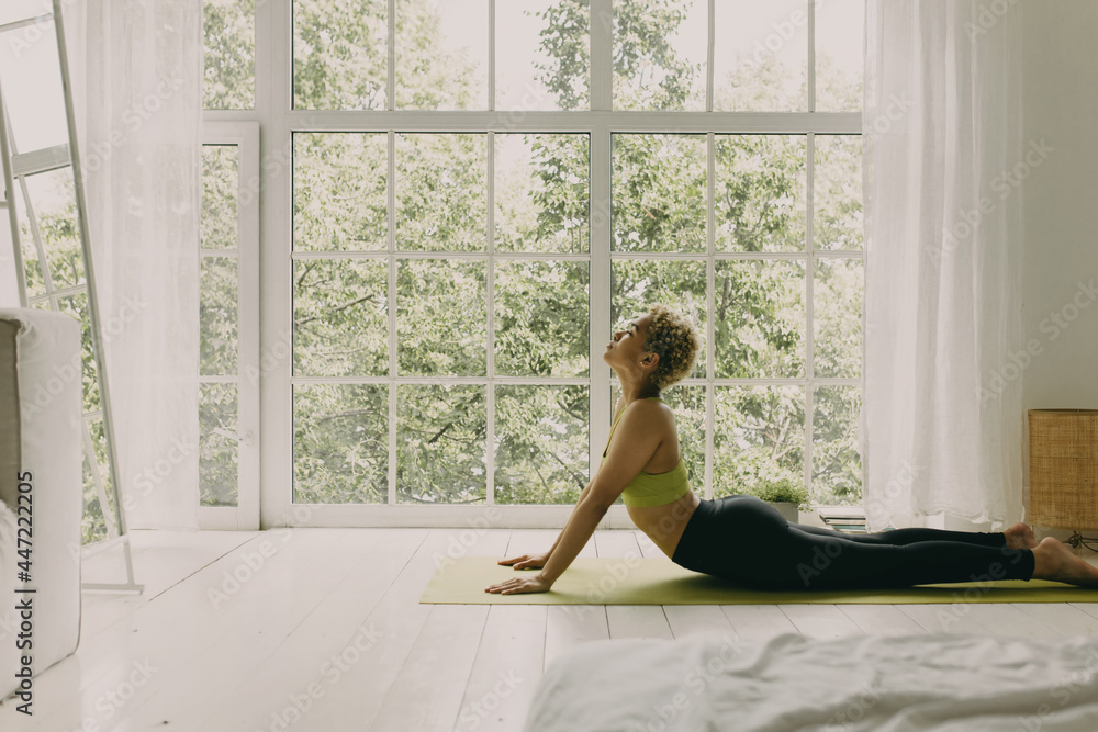 Concentrated black woman practicing yoga in light room with big windows. Position with deep breathing, morning ritual for better life. Meditation, yoga time in everyday routine concept. Copy space Concentrated black woman practicing yoga in light room with big windows. Position with deep breathing, morning ritual for better life. Meditation, yoga time in everyday routine concept. Copy space