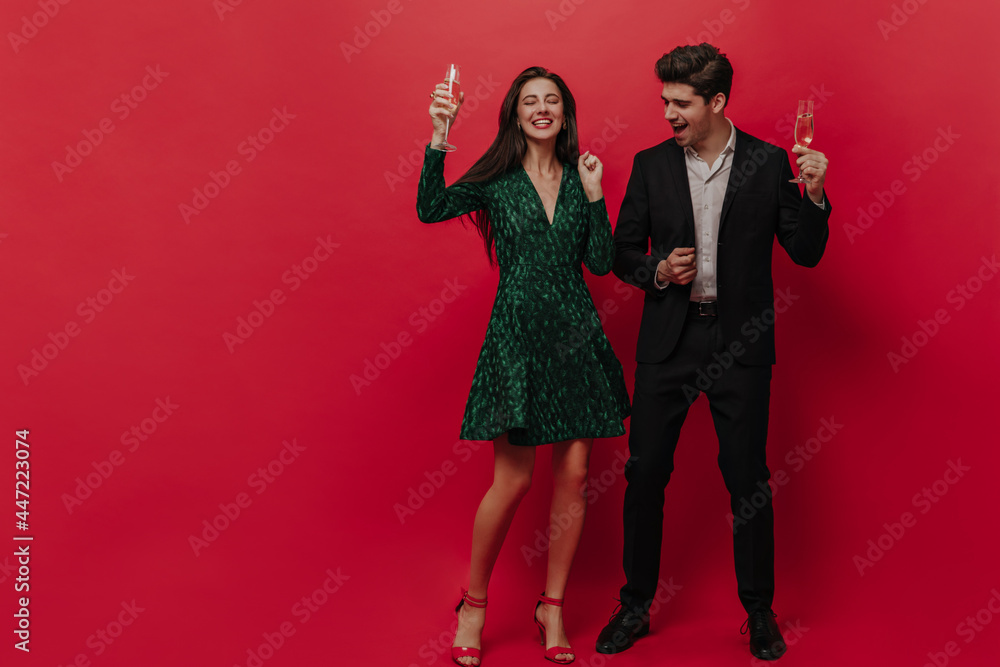 Couple of pretty people in green dress and black suit enjoying, celebrating with champagne isolated on red background Couple of pretty people in green dress and black suit enjoying, celebrating with champagne isolated on red background