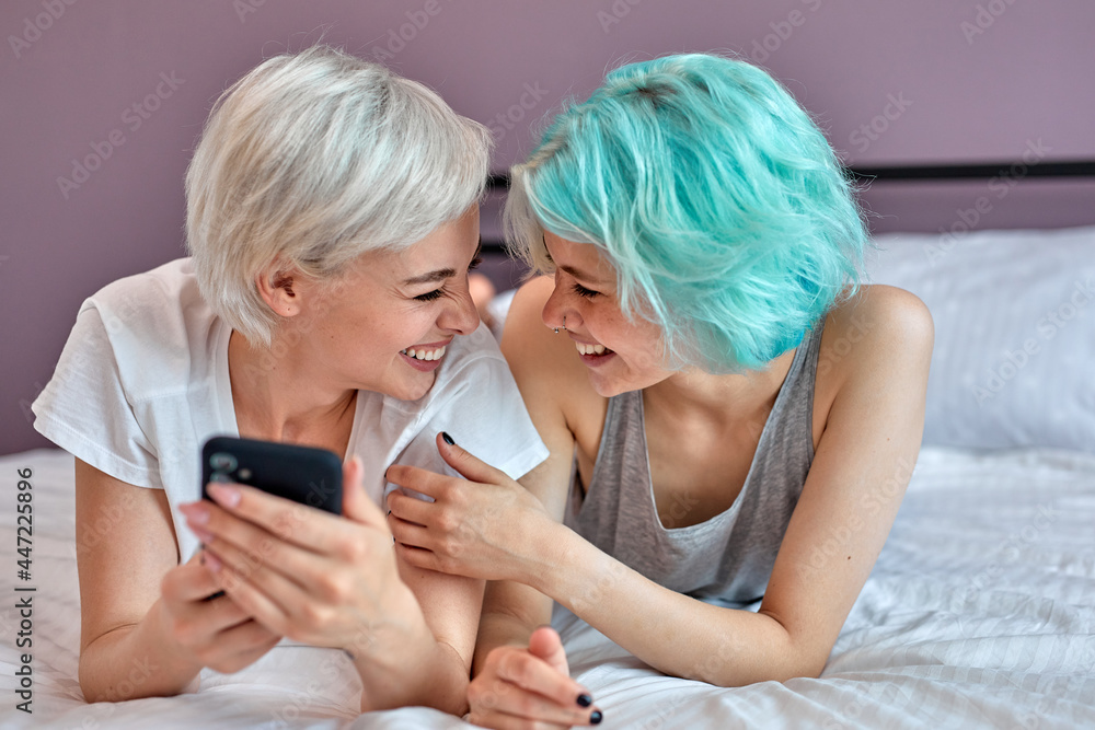 Two young attractive women lying at home on bed with smartphone, having fun, watching video film, video blog. Rest and leisure, friendship, communication, relationships. Lesbians, lgbt concept Two young attractive women lying at home on bed with smartphone, having fun, watching video film, video blog. Rest and leisure, friendship, communication, relationships. Lesbians, lgbt concept