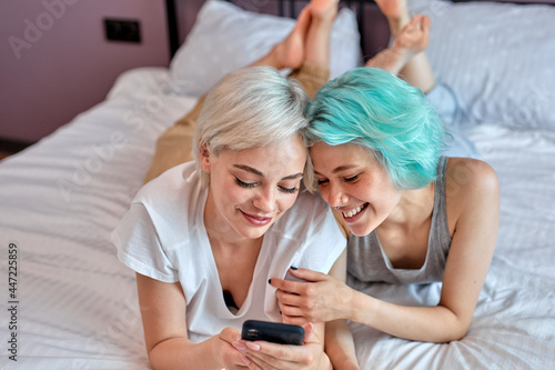 Positive lesbian couple having sun on bed using smartphone, watching interesting video.Cute Caucasian short head ladies spending nice time together at home, at weekends. Copy space, portrait