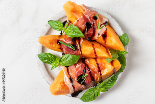 Fényképezés Melon cantaloupe slices with prosciutto ham, balsamic vinegar sauce and basil in a plate on white background