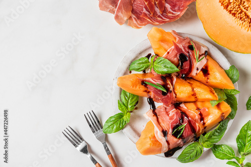 Prosciutto ham with melon cantaloupe slices, balsamic vinegar sauce and basil in a plate on white background. top view photo