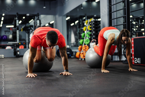 A man and a woman in sportswear do workout for the body core and abdomen. They are in a plank position with their feet on a large pilates ball while their hands are on the gym black floor. Sport, love
