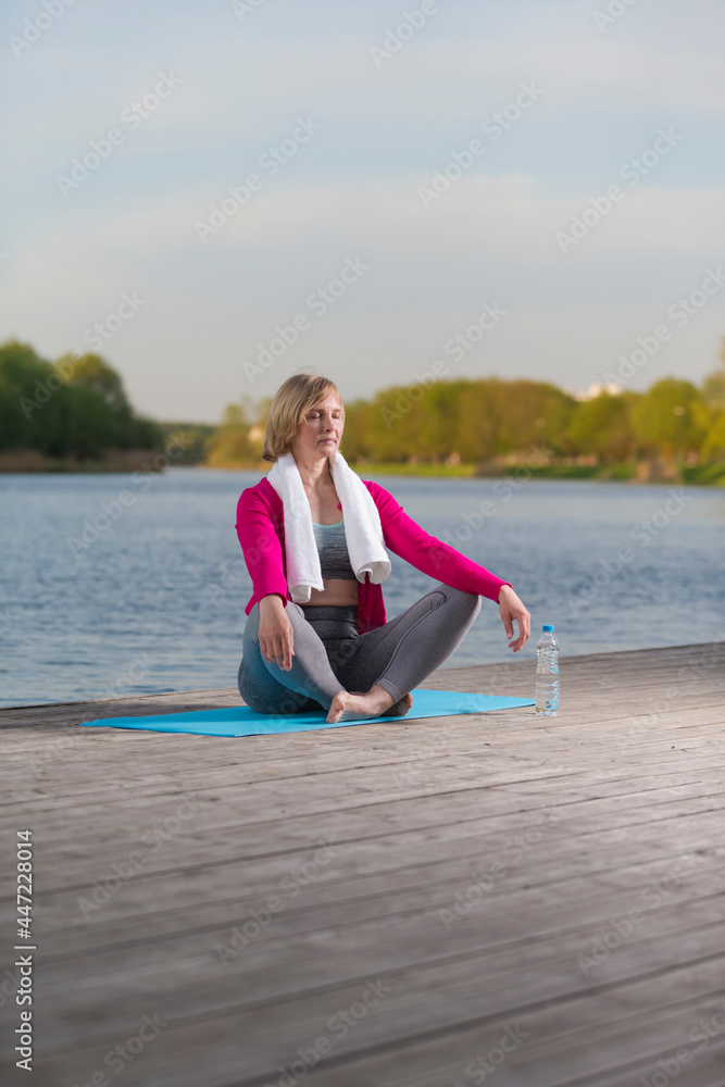 Mature Caucasian Blond Woman During Yoga Practice With Eyes Closed on Blue Mat At Water Shore Outdoor