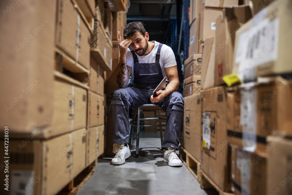 Young worried hardworking tattooed bearded blue collar worker in overalls sitting on the chair in storage of import and export firm, holding tablet and touching his head. Is delivery on time? Young worried hardworking tattooed bearded blue collar worker in overalls sitting on the chair in storage of import and export firm, holding tablet and touching his head. Is delivery on time?