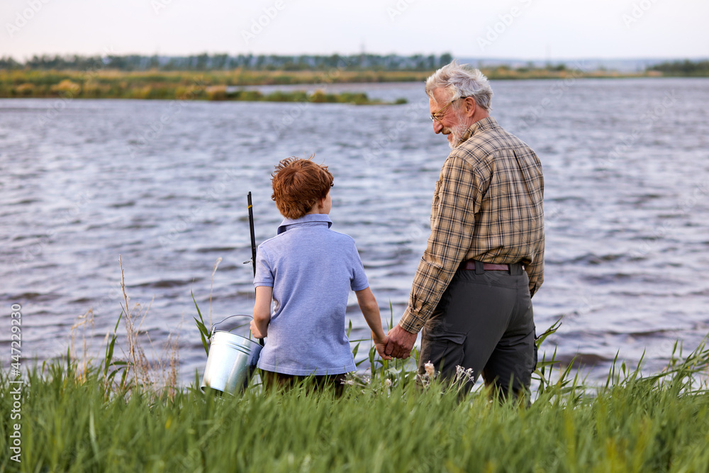 Rear View On Child boy and Grandfather in casual wear going to fish together on riverside in countryside, holdng bucket, keeping hands together. summer vacation, generation. Copy space.