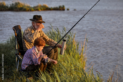Glad grey-headed man with grandson on fishing, drinking tea from thermos, have rest in nature, leisure time, enjoy and relax. Side view portrait of family in checkered shirt on lake in the evening