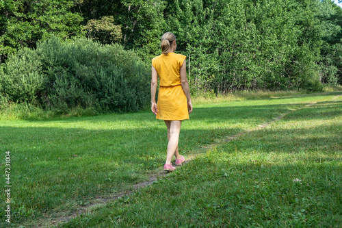 Young slender woman walks on forest pathway. Slim girl dressed in yellow short dress walking alone on the green grass trail. Healthy lifestyle fresh air. Rehabilitation after coronavirus disease.