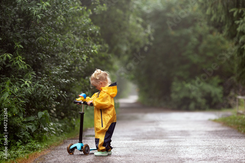 Little blonde girl wearing yellow waterproof overall standing outdoors in countryside with scooter