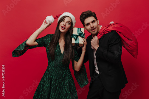 Attractive young lady in green dress and Santa hat holding gift box and posing with handsome brunette wearing black suit. Couple posing isolated on red background 
