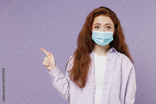 Young redhead woman 20 wear white T-shirt jacket sterile face mask ppe to safe from covid-19 flu point onworkspace area copy space mock up isolated pastel purple color wall background studio portrait
