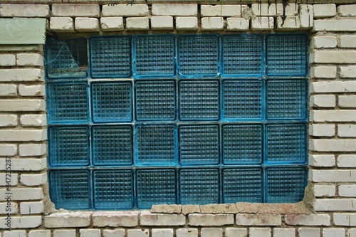 Factory warehouse window made of blue square glass framed by white brickwork wall
