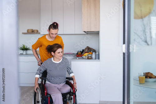Adult son helping disabled mature mother in wheelchair in kitchen indoors at home. photo