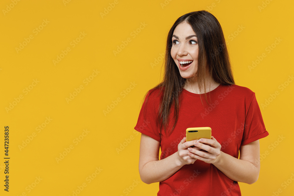 Beautiful smiling fun satisfied cheerful surprised young brunette woman 20s wears basic red t-shirt hold in hand use mobile cell phone looking aside away isolated on yellow background studio portrait Beautiful smiling fun satisfied cheerful surprised young brunette woman 20s wears basic red t-shirt hold in hand use mobile cell phone looking aside away isolated on yellow background studio portrait