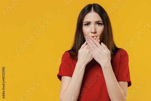 Puzzled thoughtful wistful minded sad beautiful young brunette woman 20s wears basic red t-shirt cover mouth with hand isolated on yellow background studio portrait. People emotions lifestyle concept.