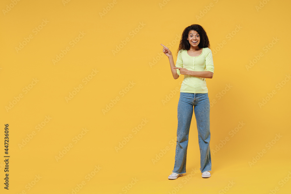 Full size body length smiling excited fun amazed african american young woman 20s wears green shirt stand pointing on workspace area copy space mock up isolated on yellow background studio portrait Full size body length smiling excited fun amazed african american young woman 20s wears green shirt stand pointing on workspace area copy space mock up isolated on yellow background studio portrait