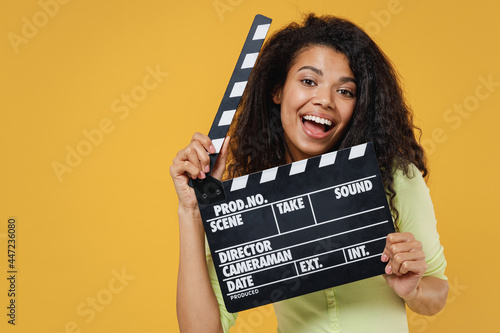 Εκτύπωση καμβά Smiling african american young woman 20s wear green shirt holding hiding behind classic black film making clapperboard isolated on yellow background studio portrait