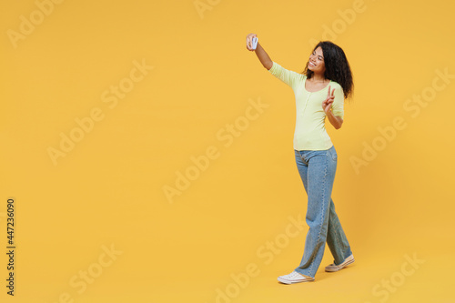 Full size body length overjoyed african american young brunette woman 20s wears green shirt go walk do selfie shot on mobile cell phone show victory sign isolated on yellow background studio portrait. © ViDi Studio
