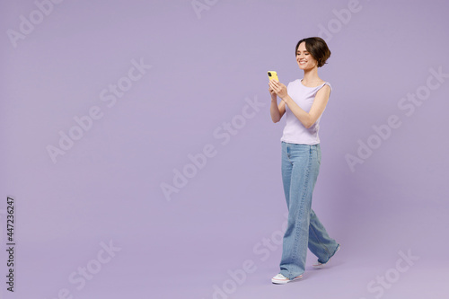 Full length side view young smiling happy fun brunette woman 20s in white t-shirt using mobile cell phone chat online browsing internet walk go isolated on pastel purple background studio portrait © ViDi Studio