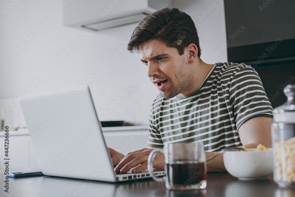 Young worried sad troubled man in striped t-shirt eat breakfast muesli cereals with milk fruit in bowl work online with laptop pc computer look for recipe cooking food in light kitchen at home alone. Young worried sad troubled man in striped t-shirt eat breakfast muesli cereals with milk fruit in bowl work online with laptop pc computer look for recipe cooking food in light kitchen at home alone.