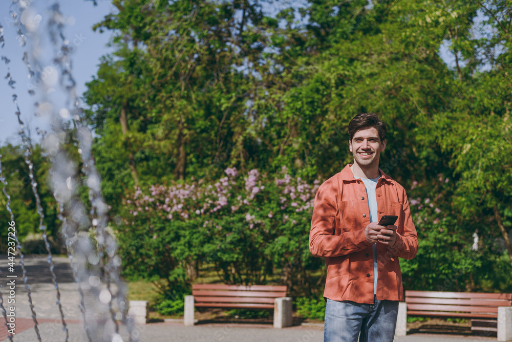 Fototapeta premium Young smiling fun man in orange jacket blue t-shirt walk using mobile cell phone near fountain rest relax in spring green city park go down alley sunshine lawn outdoor on nature Urban leisure concept