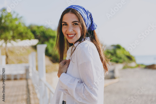 Beautiful young smiling happy traveler tourist woman 20s in blue bandana white shirt summer casual clothes standing resting outdoors at sea beach background People vacation lifestyle journey concept.