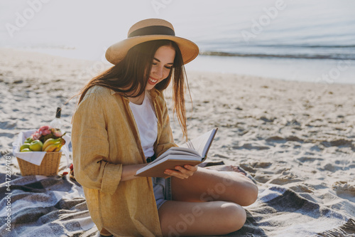 Full length young traveler tourist woman in straw hat glasses shirt summer clothes reading book sit on plaid have picnic outdoor on sea sand beach background People vacation lifestyle journey concept photo