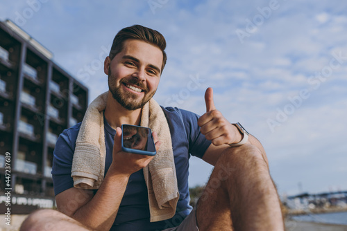 Young sporty strong fit sportsman man in sports clothes warm up training record voice message on mobile cell phoneshow thumb up at sunrise over sea beach outdoor near building in summer day morning photo