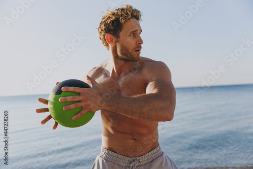 Young sporty athletic toned fit sportsman topless man 20s warm up train play hold medicine crossfit ball at sunrise sun dawn over sea beach outdoor on sand dawn beach seaside in summer day morning. photo