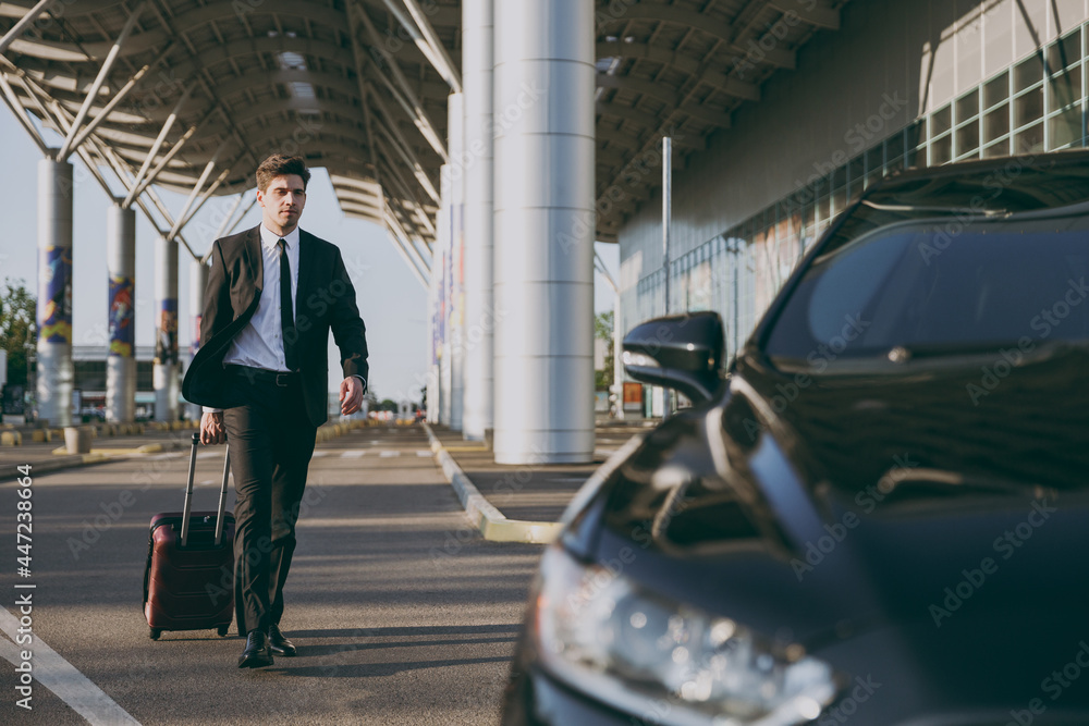 Full body young traveler businessman young man 20s wearing black dinner suit going walk outside at international airport terminal with suitcase to car booking taxi. Air flight business trip concept - obrazy, fototapety, plakaty 