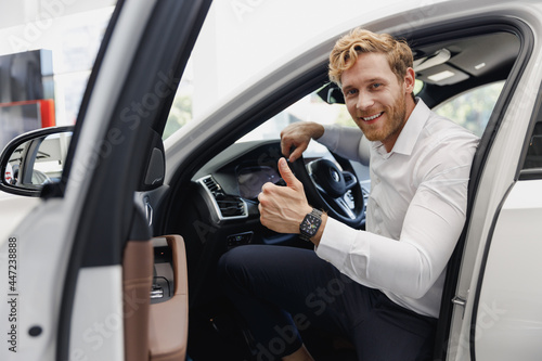Happy man customer male buyer client in shirt get out car touch door show thumb up salon drive choose auto want buy new automobile in showroom vehicle dealership store motor show indoor Sales concept.