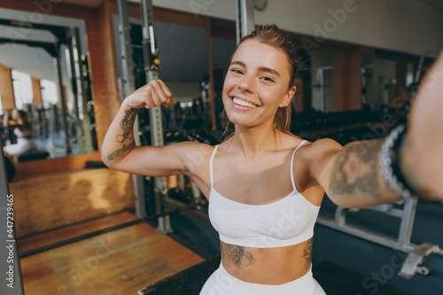 Close up young sporty athletic sportswoman woman in white sportswear warm up training sit near treadmill trainers do selfie shot on mobile cell phone show biceps in gym indoors Workout sport concept