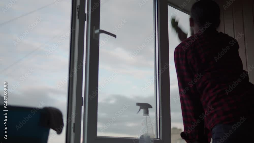 Washing window glass at home. A young long-haired man with a beard is cleaning the apartment. Sanitary worker is disinfecting panoramic windows in an empty room. purity, cleanliness, change concept Washing window glass at home. A young long-haired man with a beard is cleaning the apartment. Sanitary worker is disinfecting panoramic windows in an empty room. purity, cleanliness, change concept
