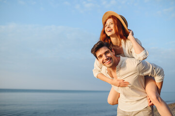 Young smiling happy couple two friends family man woman in white clothes boyfriend give piggyback ride to joyful, girlfriend sit on back at sunrise over sea beach ocean outdoor seaside in summer day.