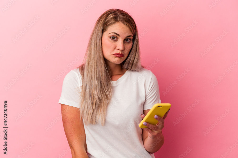 Young russian woman holding a mobile phone isolated on pink background shrugs shoulders and open eyes confused. Young russian woman holding a mobile phone isolated on pink background shrugs shoulders and open eyes confused.
